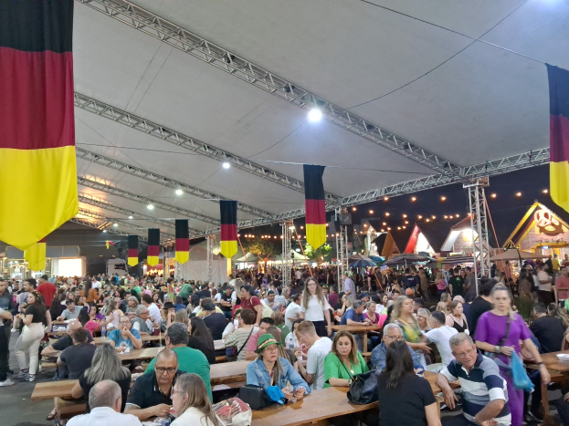 Eine große Gruppe von Menschen sitzt an langen Tischen unter einem Zelt auf dem Oktoberfest in München, Deutschland, während einige in der Nähe stehen und Taschen auf den Tischen liegen. Im Hintergrund sind Zelte, Lichter und Fahnen zu sehen.