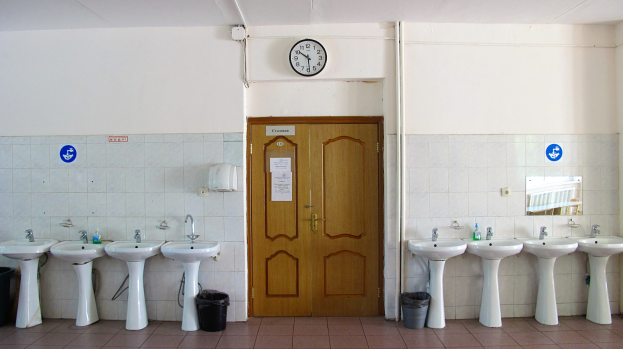 Ein Badezimmer mit mehreren Waschbecken, bodenebenen Müllbehältern, einer Tür im Hintergrund, einer Wanduhr und einem Spiegel auf der rechten Seite.