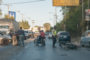 Eine Gruppe von Menschen um ein verunglücktes Motorrad auf der Straße herumsteht, umgeben von mehreren Fahrzeugen, einschließlich eines Lastwagens, und einem Hintergrund aus Bäumen, Pfählen, Lampen und Schildern.