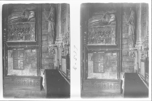 Ein Schwarz-Weiß-Foto einer Grabkammer mit einer hölzernen Wand, die mit Schnitzereien und Skulpturen verziert ist und in der Mitte einen Sarg und im Vordergrund einen Tisch mit einer Blumenvase zeigt.