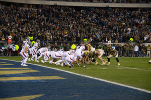Ein Fußballspiel im Gange mit Spielern in Sportkleidung und Helmen auf dem Feld, betrachtet von einem Stadionplatz.