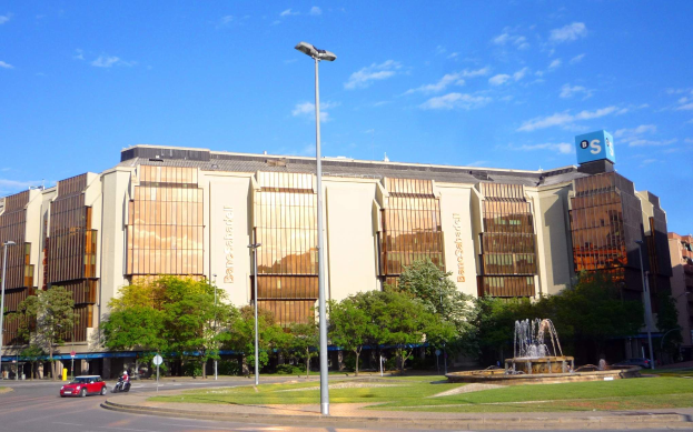 Bancroft-Stadion, ein großes Gebäude mit einem Brunnen davor, umgeben von Straßenmasten, Straßenlaternen, Kraftfahrzeugen, Fußgängern, Bäumen und einem bewölkten Himmel.